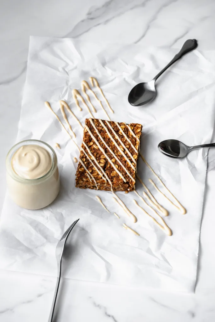 Barre gâteau aux carottes recouverte d'un filet de glaçage crémeux à la vanille avec des ustensiles et un pot de glaçage à côté